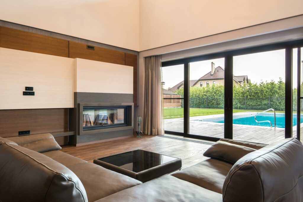 Modern living room with a leather sectional sofa, fireplace, and large sliding glass doors opening to a patio and outdoor swimming pool.